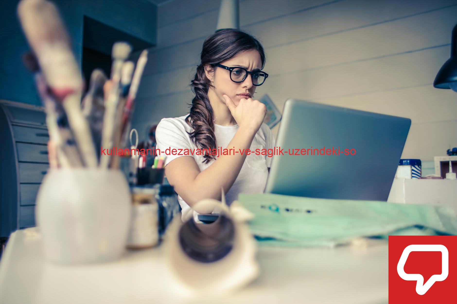 Young woman with glasses deeply focused on a laptop surrounded by art supplies in a home office.