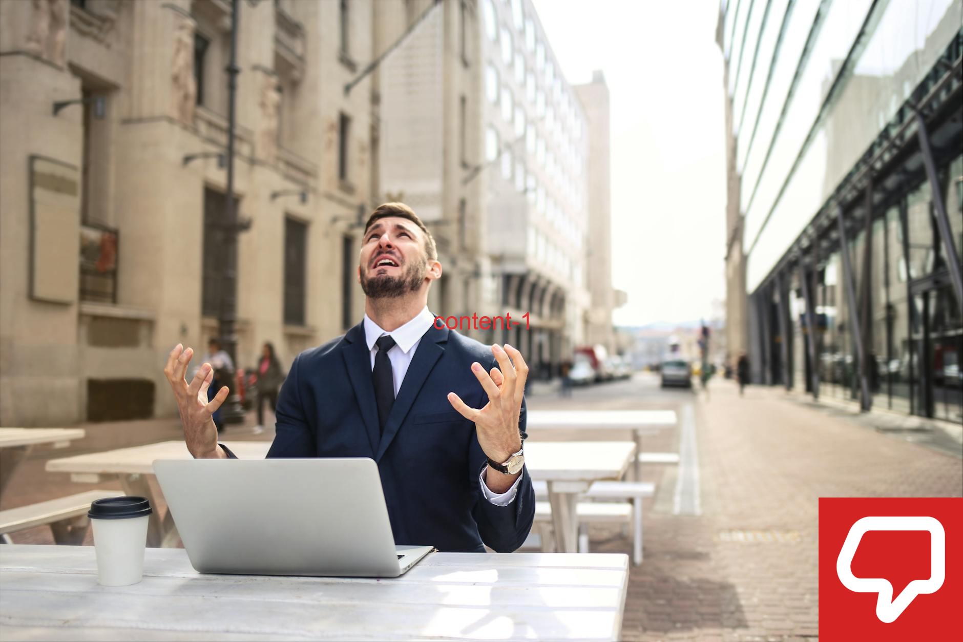 Businessman in suit showing frustration at outdoor table with laptop and coffee.