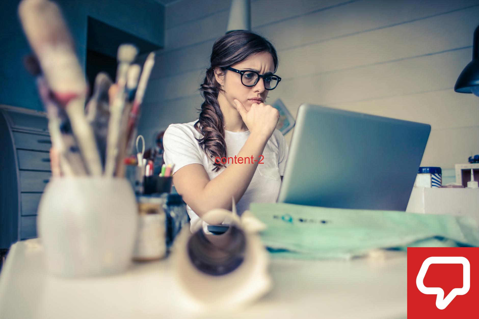 Young woman with glasses deeply focused on a laptop surrounded by art supplies in a home office.