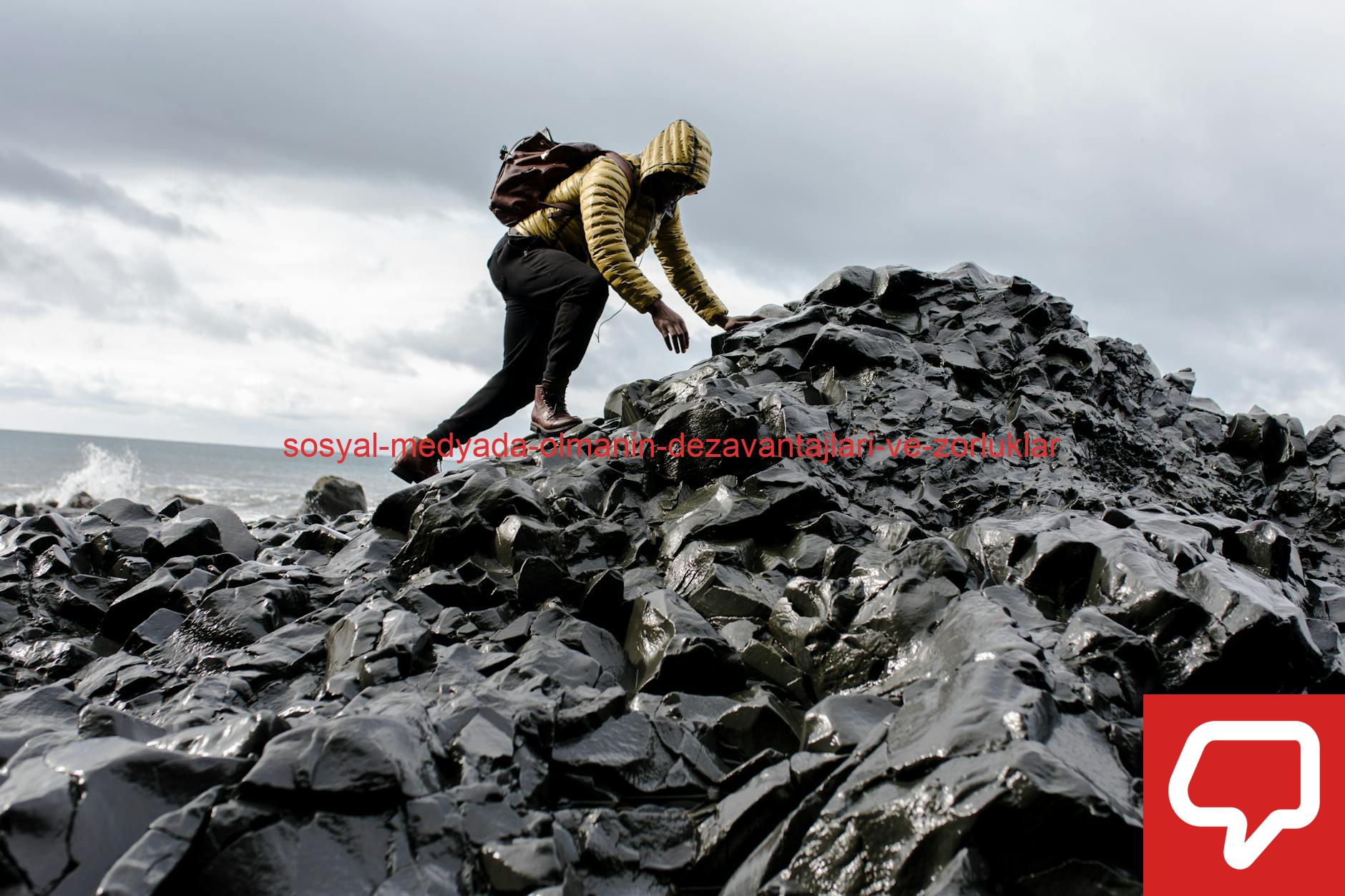 Person in yellow jacket climbs black rocky terrain by the ocean under cloudy skies.